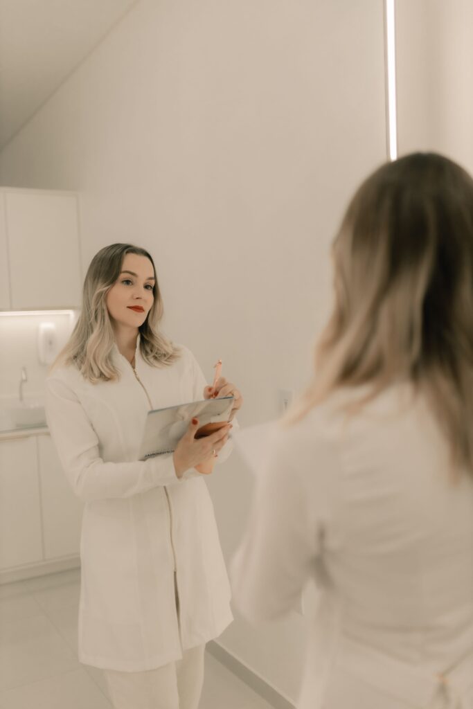 A woman in white attire holds a tablet while looking in the mirror.